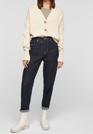 Béžový pletený kardigan s velkými knoflíky, v kombinaci s tmavě modrými džíny s vysokým pasem a bílými kotníkovými botami s texturovaným povrchem.