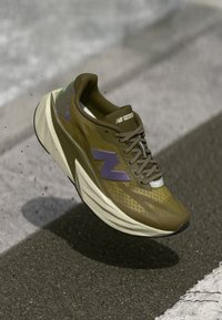 Zapatilla de correr verde y marrón con parte superior de malla, acentos morados y suela gruesa y acolchada, que cuenta con detalles texturizados y cordones elásticos.
