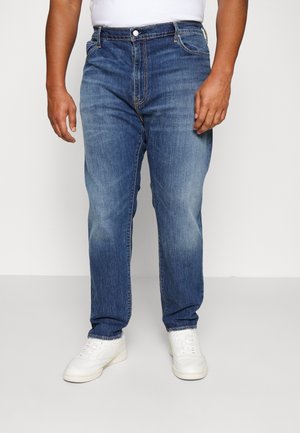 Mand iført blå, falmede jeans med lige ben og hvide sneakers, stående mod en ensartet lys baggrund.