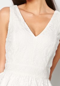 Abito in pizzo bianco con scollatura a V e motivi floreali. Il tessuto testurizzato presenta una finitura morbida e stratificata con accenti arricciati all'orlo.