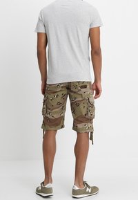 Pantalones cortos de camuflaje en tono beige con manchas negras y marrones. Presentan grandes bolsillos laterales y un dobladillo enrollado. Combinados con una camiseta gris ajustada y zapatillas.