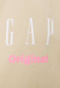 Camiseta de algodón beige con letras grandes en blanco que dicen "GAP" y "Original" en rosa neón, con un acabado de tela lisa y sin textura.