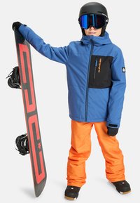 Enfant portant une veste bleue et noire et un pantalon orange, avec un casque de ski et des lunettes de ski, tenant verticalement un snowboard noir avec un design rouge.