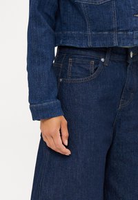 Donkerblauwe denimjacket met een cropped ontwerp, met een rechte kraag en zichtbare stiksels. Hand rustend op donkere denim broek.