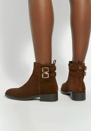Brown suede ankle boots with a low heel, featuring two gold-tone buckles and a rear zipper; smooth texture with rounded toe design.