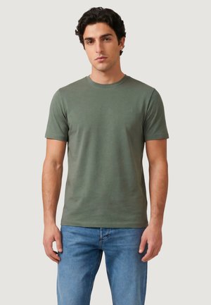 Giovane uomo che indossa una t-shirt a maniche corte verde oliva e jeans blu, in piedi davanti a uno sfondo chiaro e uniforme.