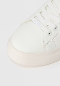 Nahaufnahme eines weißen Sneakers mit dicker Sohle und weißen Schnürsenkeln, der an einem Schnürsenkel ein kleines "GUESS"-Tag hat.