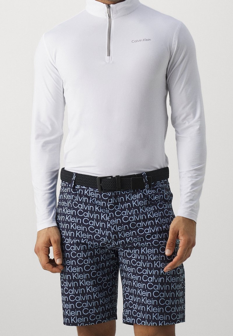 Hombre vestido con una camiseta de manga larga blanca con cremallera de un cuarto y pantalones cortos azul marino con texto repetido en blanco "Calvin Klein", asegurados con un cinturón negro.