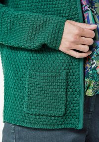 Groene gebreide cardigan met een textuurpatroon, ribbelmanchetten en een voorzak. Met rits sluiting en gedragen over een patroon shirt.