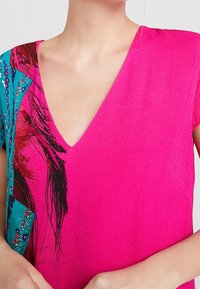 Rosa V-Ausschnitt-Oberteil mit kurzen Ärmeln, das auf einer Seite Akzente mit türkisfarbenem Muster, Verzierungen und feinen strukturierten Details aufweist.