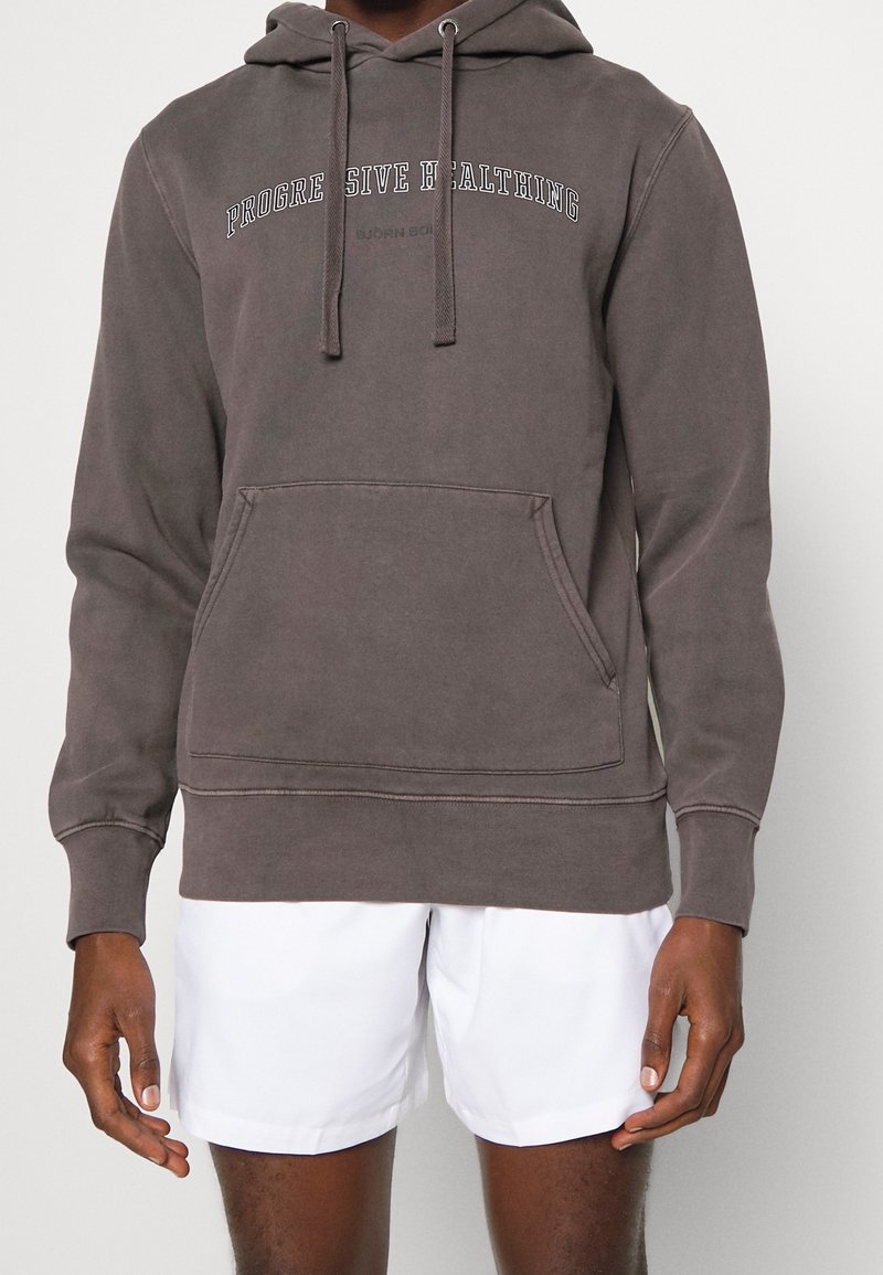 Sweat à capuche gris foncé avec inscription "PROGRESSIVE HEALTHING" et poche kangourou, porté avec un short blanc sur une personne à la peau foncée.