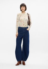 Μάλλινη μπλούζα με μακριά μανίκια, εφαρμοστή, με λουλουδάτο και γεωμετρικό σχέδιο σε κρεμ χρώμα, συνδυασμένη με ανοιχτά μπλε παντελόνια με φαρδιά πόδια. Καφέ τσάντα ώμου και μπότες με τακούνια.