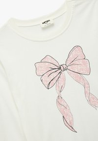 Hvid bomulds T-shirt med en lyserød omrids-tegning af en sløjfe med indviklede mønstre, rund halsudskæring og korte ærmer.