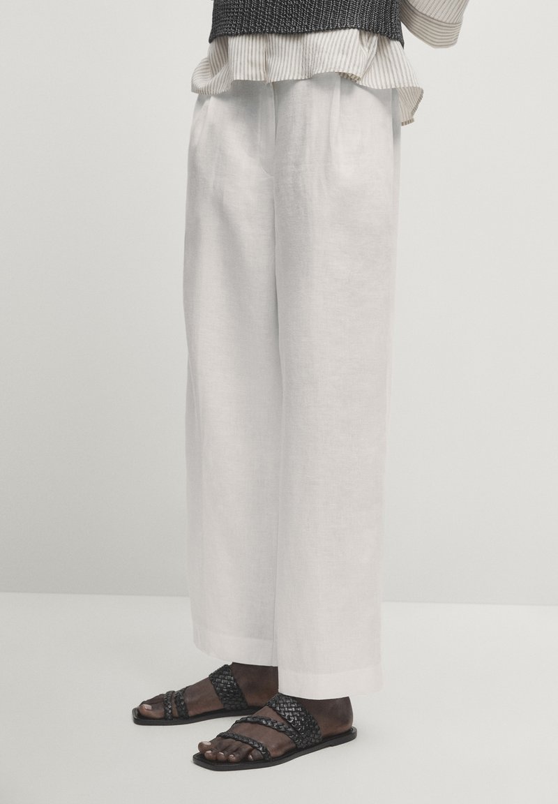 Witte linnen wijde broek met een relaxte fit, gecombineerd met zwarte gevlochten sandalen met meerdere banden. De textuur lijkt licht en luchtig.