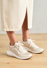 Beige sportieve sneakers met een gestructureerde bovenkant, vetersluiting, zijrits en een witte plateauzool met metallic accenten.
