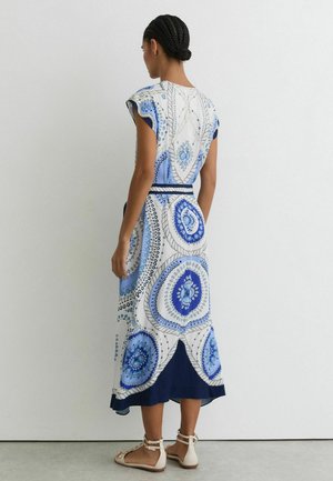 Žena s vrkočmi stojí chrbtom, oblečená v bezrukávové, na členky dlhej šaty s modrobielymi kruhovými vzormi a bielych sandáloch.