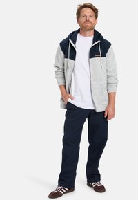 Sudadera con capucha en bloques de color gris y marino, con cremallera, bolsillos frontales y cordón ajustable, combinada con pantalones marinos y zapatillas marrones.