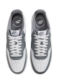 Nike Sneakers mit einem weißen Leder-Obermaterial und grauen Akzenten. Zu den Merkmalen gehören durchbrochene Muster, flache weiße Schnürsenkel und eine Markenkennzeichnung auf der Zunge.