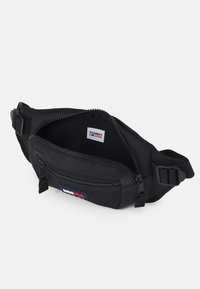 Porte-monnaie noir en tissu avec un compartiment principal zippé, une poche avant, une sangle réglable et un patch logo Tommy Jeans en rouge, blanc et bleu.