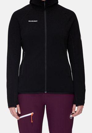 Czarna kurtka zapinana na zamek z kapturem i logo Mammut, noszona z ciemnofioletowymi spodniami z kieszenią na zamek i logo na udzie.