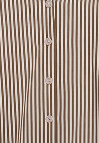 Brun- och vitrandig blus med knäppning fram. Har fem runda, iriserande knappar längs mitten och mjuk, slät tygstruktur.