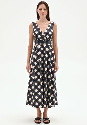 Femme portant une robe midi sans manches à carreaux noirs et blancs avec un profond décolleté en V et une large ceinture, debout sur fond blanc.