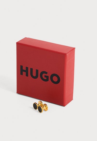 Paire de boutons de manchette en or avec des inserts circulaires noirs placés à côté d'une boîte rouge étiquetée HUGO sur une surface blanche.