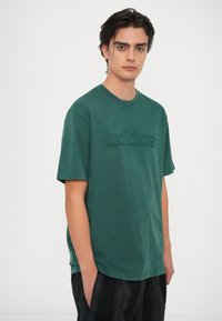 Zelené bavlnené tričko s krátkymi rukávmi, okrúhlym výstrihom a textúrovaným logom "adidas" v mierne tmavšej zelenej farbe. Hladký povrch, uvoľnený strih.