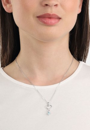 Collana a forma di cuore in argento con un design intrecciato, caratterizzata da un ciondolo di pietra preziosa blu chiaro. Indossata sopra una maglietta bianca a coste.
