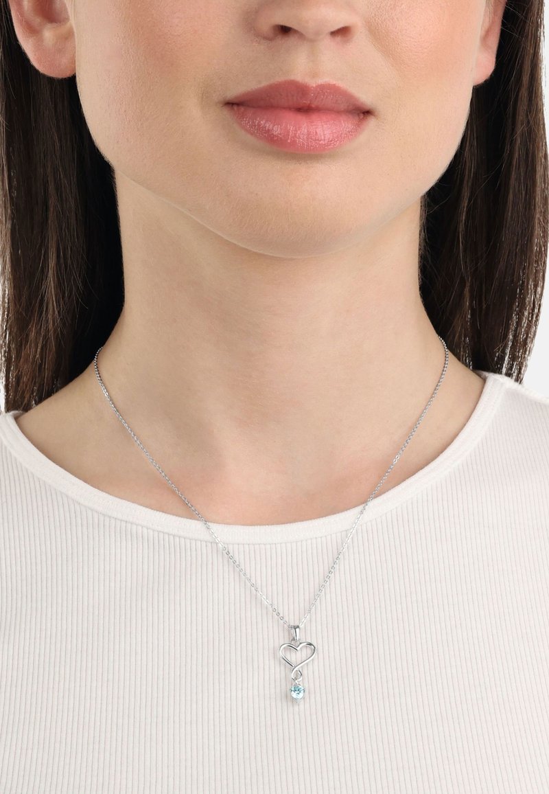 Silver hängande halsband i form av ett hjärta med en sammanflätad design, prydd med en ljusblå ädelsten i droppeform. Bärs ovanpå en vit ribbad topp.