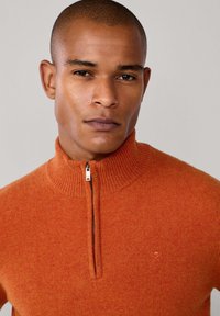 Maglione arancione con zip e collo a costine, realizzato in un materiale morbido. Presenta un piccolo logo sul petto. Texture liscia e colore uniforme.