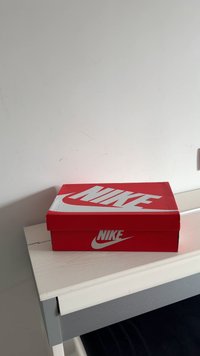 Caixa de sapatos vermelha da Nike com um logotipo branco na tampa, de formato retangular, feita de papelão. Posicionada sobre uma superfície de cor clara.