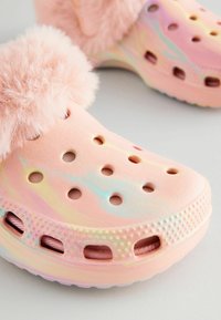 Sabots roses duveteux avec une finition brillante et irisée. Dotés de trous de ventilation et d'une semelle texturée. Un bord en fourrure douce en haut ajoute du détail.