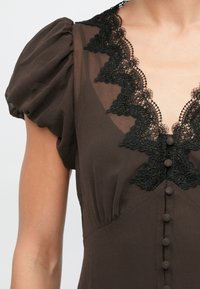 Καφέ φόρεμα με κοντά φουσκωτά μανίκια, διαθέτει λαιμόκοψη με μαύρη δαντέλα και κλείσιμο με κουμπιά μπροστά, με λείο ύφασμα.