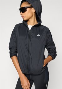 Μαύρο ελαφρύ μπουφάν με κουκούλα, φερμουάρ μπροστά και λογότυπο. Διαθέτει ελαστικά μανίκια και λεία υφή. Φοριέται με μαύρα κολάν.