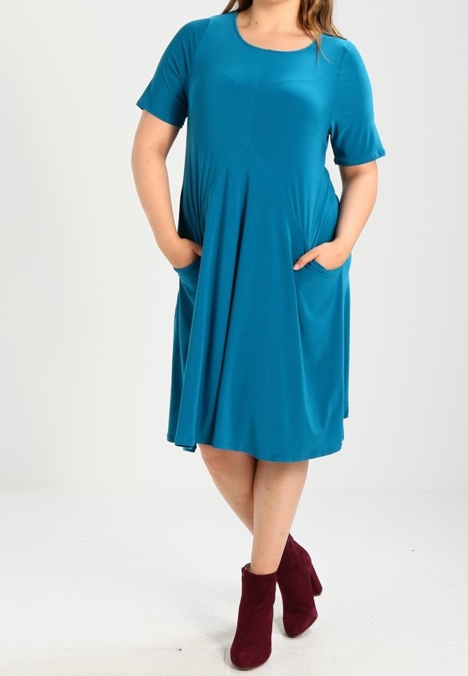 Femme portant une robe turquoise longueur genoux à manches courtes, les mains dans les poches, et des bottines bordeaux, debout, jambes croisées.