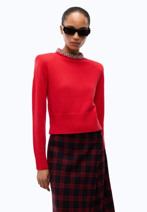 Donna che indossa occhiali da sole rettangolari neri, un maglione rosso con scollo gioiello e una gonna a quadri nera e rossa su sfondo bianco.