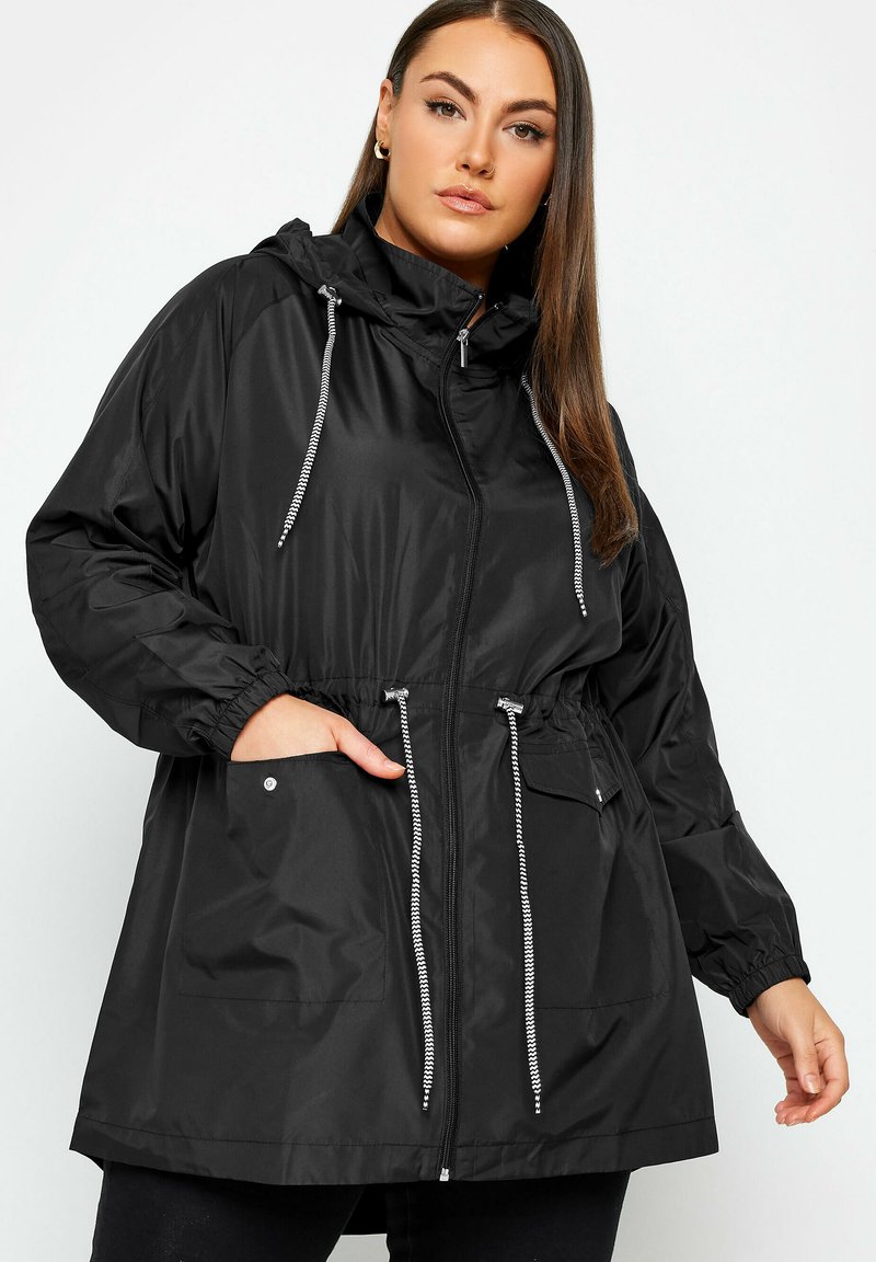 Vrouw die een zwarte regenjas met capuchon draagt, voorzien van een ritssluiting aan de voorkant, trekkoorden, elastische manchetten en grote voorzakken, staand tegen een witte achtergrond.