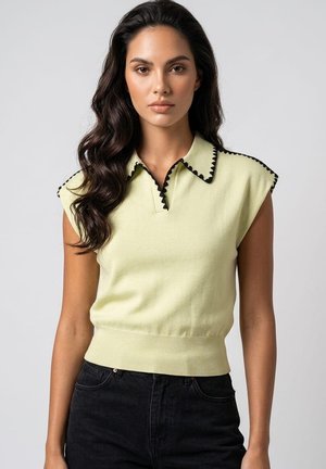 Mujer con cabello largo y oscuro lleva una camiseta de punto sin mangas de color amarillo pálido con ribete negro y vaqueros negros de talle alto sobre un fondo liso.