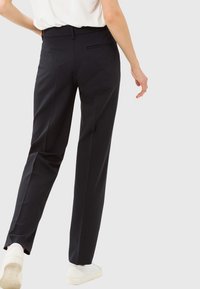 Osoba w czarnych, dopasowanych spodniach z detalem tylniej kieszeni oraz białych trampkach, idąca na tle jednolitego, jasnego tła.