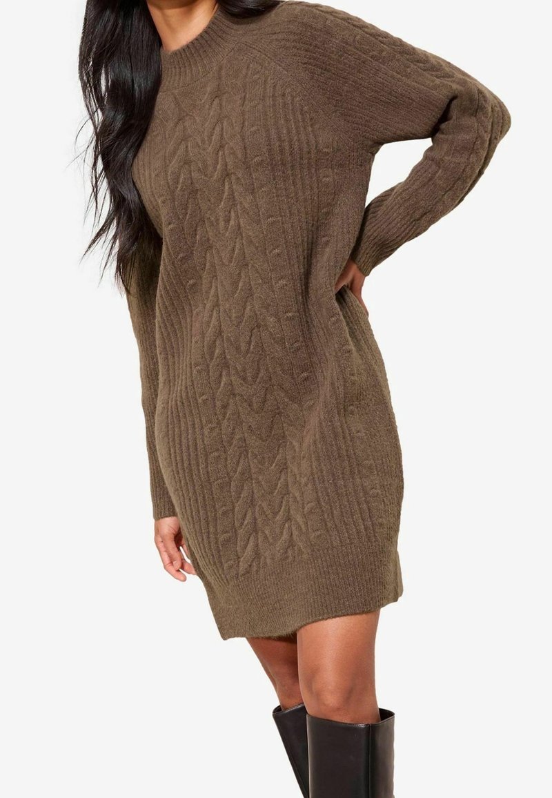 Femme portant une robe pull en maille torsadée marron et des bottes noires montant jusqu'aux genoux, posant avec une main sur la hanche sur un fond blanc.