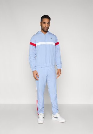 Man staat en draagt een lichtblauw trainingspak met rode en witte strepen, witte sneakers, tegen een effen lichte achtergrond.