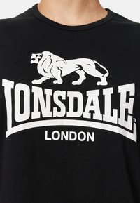Juodas medvilnės marškinėliai su baltu liūto grafiniu atvaizdu ir „LONSDALE“ tekstu stambiu didžiosiomis raidėmis. Klasikinis apvalios iškirptės dizainas su trumomis rankovėmis.
