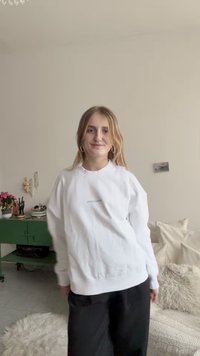 Hvit genser med rund hals og avslappet passform, med en liten grå logodetalj. Brukt med svarte, løstsittende bukser.