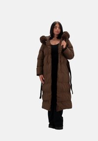 Cappotto imbottito marrone con cappuccio rifinito in pelliccia, lunghezza lunga, design trapuntato, chiusura frontale e accentuato da una cintura annodata. Indossato sopra un completo nero.