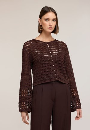 Kobieta w ciemnobrązowym, szydełkowanym rozpinanym kardiganie z długimi rękawami i dopasowanych brązowych spodniach, stojąca na tle jednolitego tła.