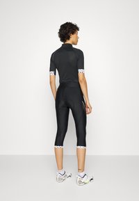 Camisola de ciclismo preta com mangas curtas em malha, apresentando detalhes em bolinhas. Calções capri pretos ajustados com um design elegante. Sapatos brancos e com padrão.