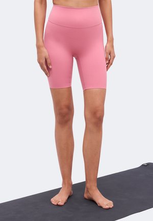 Γυναίκα στέκεται ξυπόλυτη σε μαύρο στρώμα γιόγκα φορώντας ροζ ποδηλατικό σορτς με ψηλή μέση σε απλό φόντο.