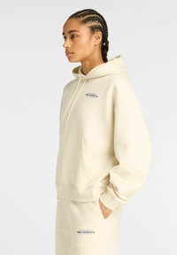 Femme aux cheveux tressés portant un sweat à capuche crème de New Balance et un pantalon de survêtement assorti, se tenant avec une main dans la poche, tournant à gauche.