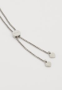 Metal necklace with a central disc featuring "LIU•JO" logo, two chains ending in silver heart-shaped pendants, textured links, and shiny finish.